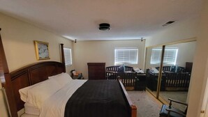 Une chambre avec un lit en bois, une commode, une chaise et un berceau. Il y a deux fenêtres avec des stores, une bouche d’aération au plafond et un miroir sur le mur.
