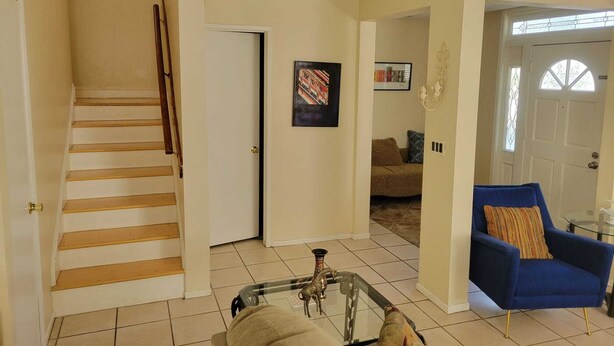 Un salon avec un escalier, un fauteuil bleu, une table basse en verre et une porte donnant sur une autre pièce.