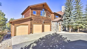 Cozy cabin-style home with a garage entrance, stone chimney, and sloped roof, set amidst mature evergreen trees.