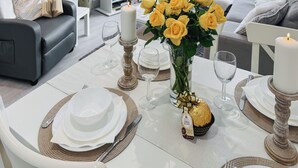 Comedor con una mesa blanca dispuesta para cuatro, con platos de borde dorado, cristalería transparente y un centro de mesa con rosas amarillas en un florero de vidrio.