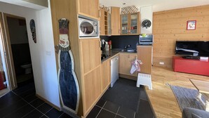 Cocina compacta con armarios de madera, microondas, pileta y un refrigerador pequeño. Hay un sofá rojo y un televisor en la sala de estar.