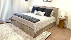 Une chambre avec un grand lit, une table de chevet avec une lampe, une fenêtre avec des stores et un tapis.