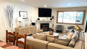 Un salon comprenant un canapé, une table basse et une cheminée. Un téléviseur est fixé au-dessus de la cheminée, et il y a une fenêtre avec des stores.