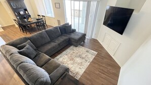 A living room with a sectional sofa, a flat-screen TV mounted on the wall, and a dining area with a table and chairs. - Beautiful, cozy home minutes from Laughlin and the river!! (Bullhead City)