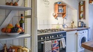 A kitchen with a gas stove, oven, and washing machine, shelves with fruits and vegetables, and a wooden shelf with kitchen items. - Hoopers Farmhouse - UK36307 (Tilshead)