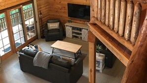 Ein gemütliches Blockhaus-Interieur mit Schlafzimmer-Galerie, einem Wohnzimmer inklusive Sofa und Fernseher sowie einem Balkon mit Geländer.