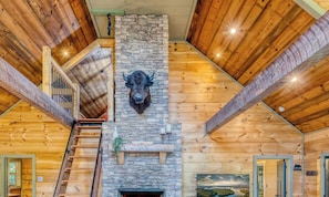 A log cabin interior with a stone fireplace, wooden beams, a staircase, and a bull head mounted on the wall.