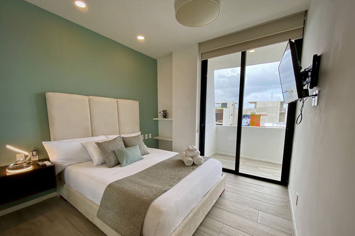 Une chambre d’hôtel moderne dotée d’un grand lit, d’une table de chevet avec une lampe, d’un téléviseur fixé au mur et d’un balcon avec vue.