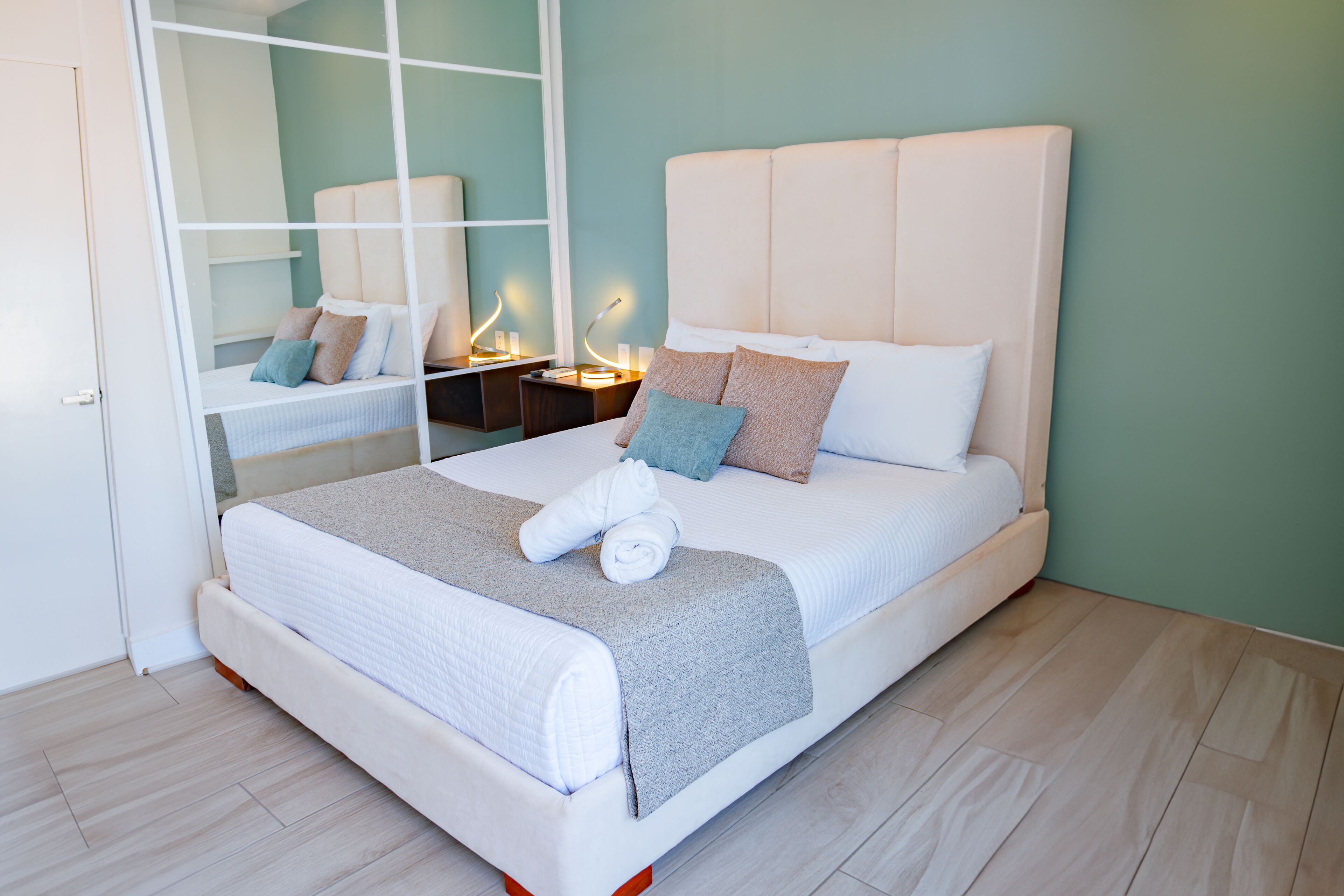 A neatly made bed with a white headboard, a grey blanket, and several pillows. There is a small bedside table with a lamp and a mirror. The room has light wooden flooring and teal walls.