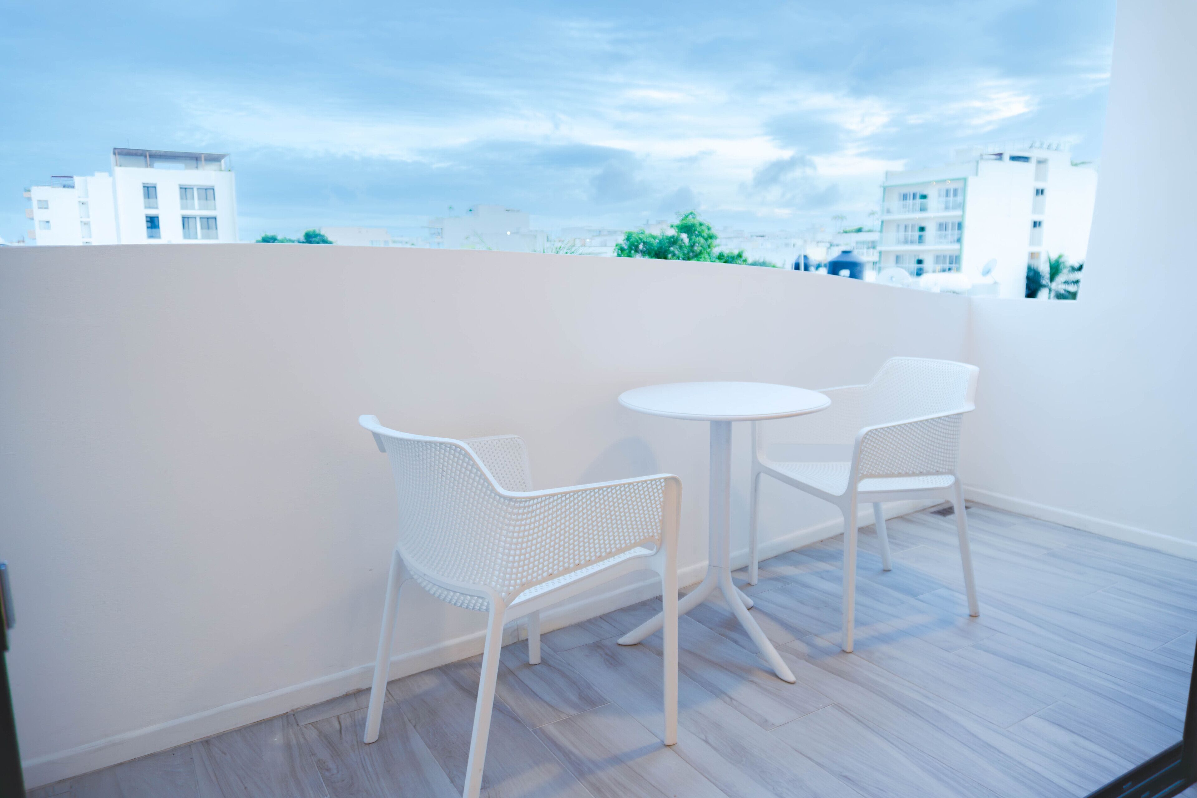 Ein Balkon mit zwei weißen Stühlen und einem kleinen Tisch, mit Blick auf eine Stadtlandschaft mit modernen Gebäuden.