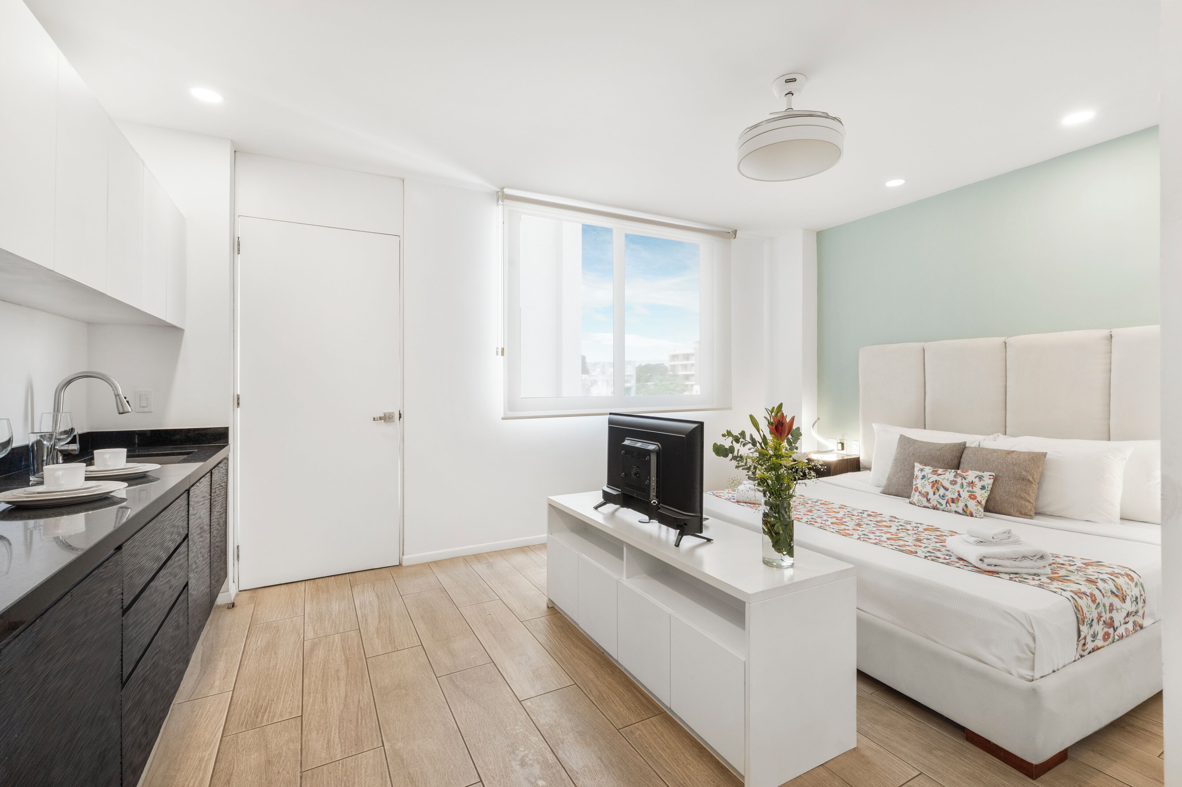 A modern hotel room with a kitchenette, a bed with a floral bedspread, a large window, and a ceiling fan.