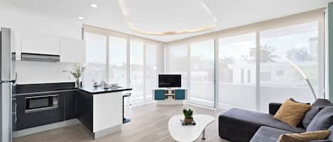A modern kitchen with a flat-screen TV, a dining area with a white table and a sofa, and large windows offering a city view.
