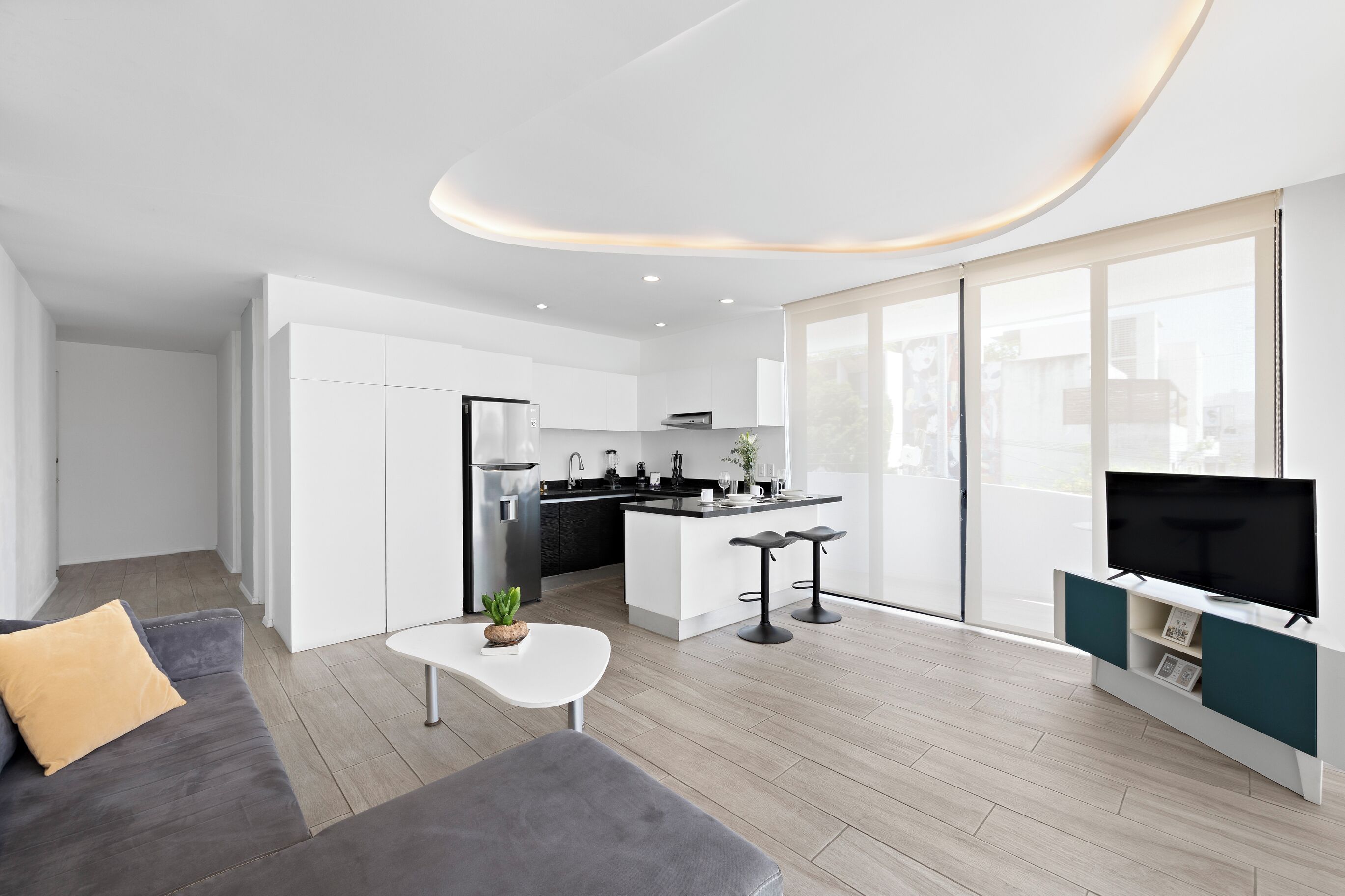 A modern living room with a grey sofa, a white coffee table, a kitchen area with white cabinets, and a flat-screen TV on a stand.