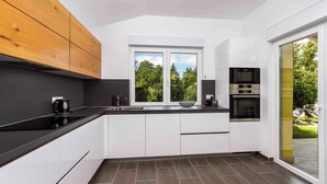 Een moderne keuken met een zwart aanrecht, witte kasten en een houten bovenkastje. Er is een raam met uitzicht op bomen en een schuifdeur naar een buitenruimte.