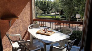 Un balcon avec une table et des chaises blanches, un plateau avec des verres et une carafe, et une vue sur un jardin et une rue.
