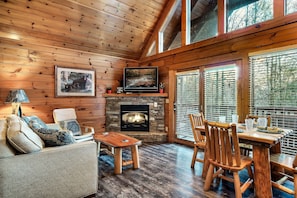 A cozy cabin interior with a stone fireplace, wooden furniture, and large windows. - Seasonal Outdoor Pool 🐻 Family Friendly🍂Cozy Cabin 🏔3 mi to Strip 🍁Hot Tub (Gatlinburg)