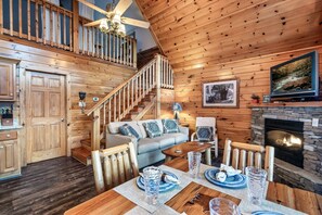A wooden interior with a staircase, a fireplace, a sofa, and a dining table set for four. - Seasonal Outdoor Pool 🐻 Family Friendly🍂Cozy Cabin 🏔3 mi to Strip 🍁Hot Tub (Gatlinburg)