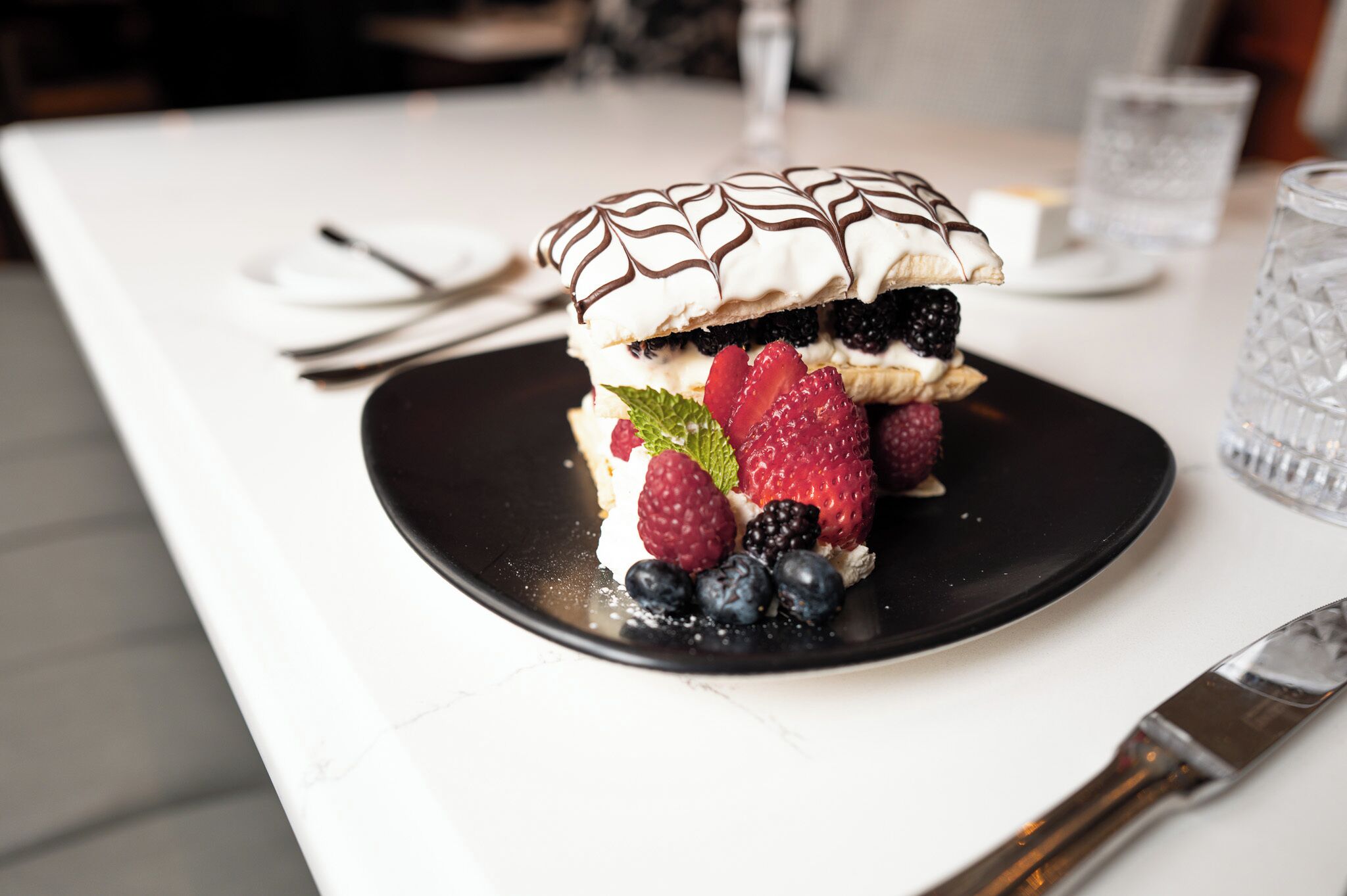 Un dessert servi sur une assiette noire, composé de fraises, de myrtilles et d’une feuille de menthe, le tout posé sur une table blanche.