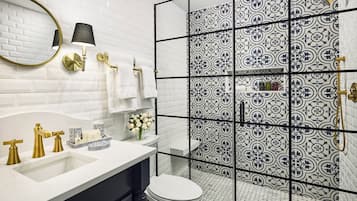 Et badeværelse med en hvid vask, guldfarvede armaturer, et toilet, en bruseniche med sorte og hvide fliser samt et spejl med gylden ramme.