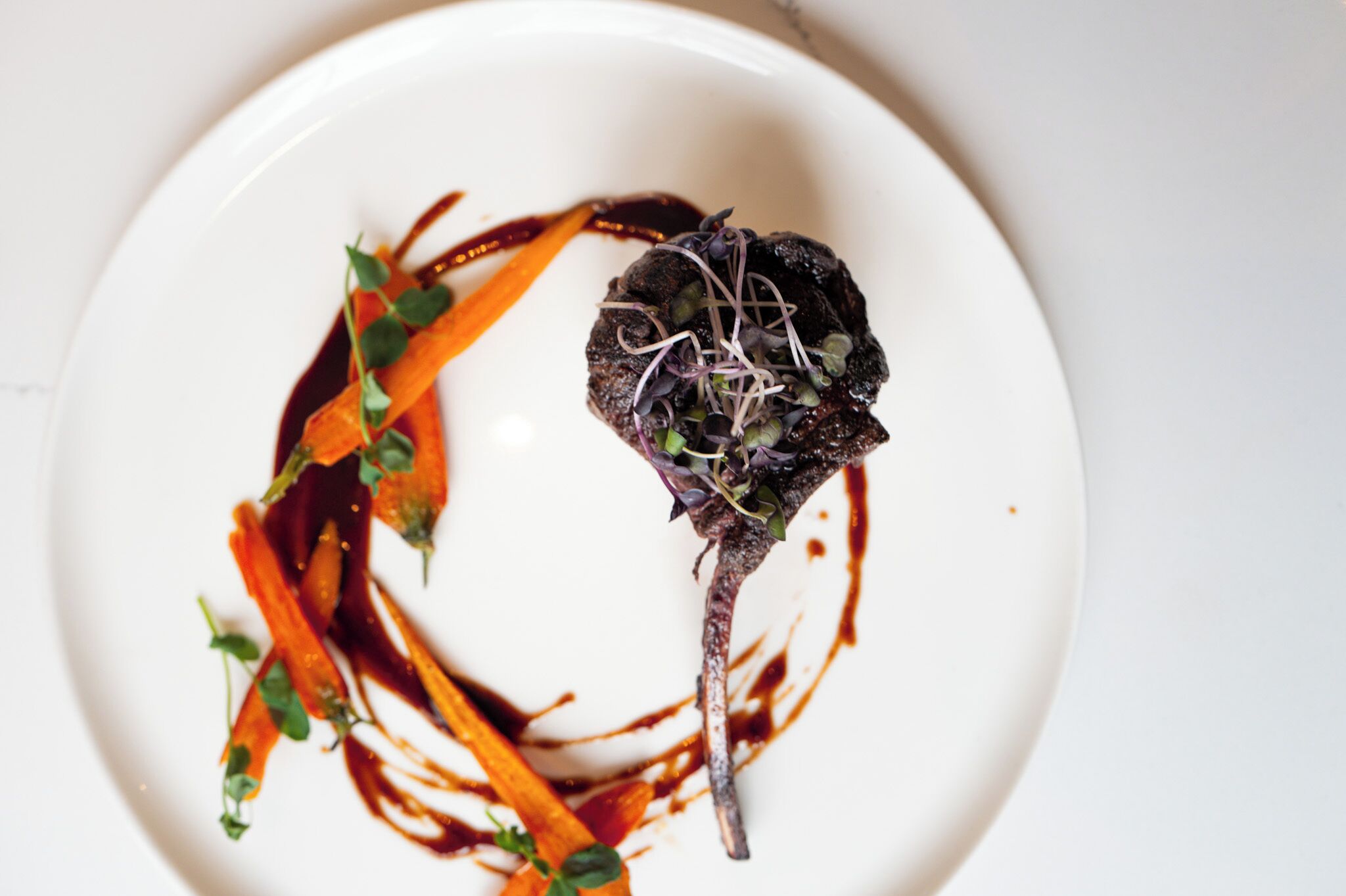 Une assiette blanche avec une portion de viande sombre en plat principal, entourée de lanières de carottes et de jeunes pousses, et nappée d’une sauce foncée.