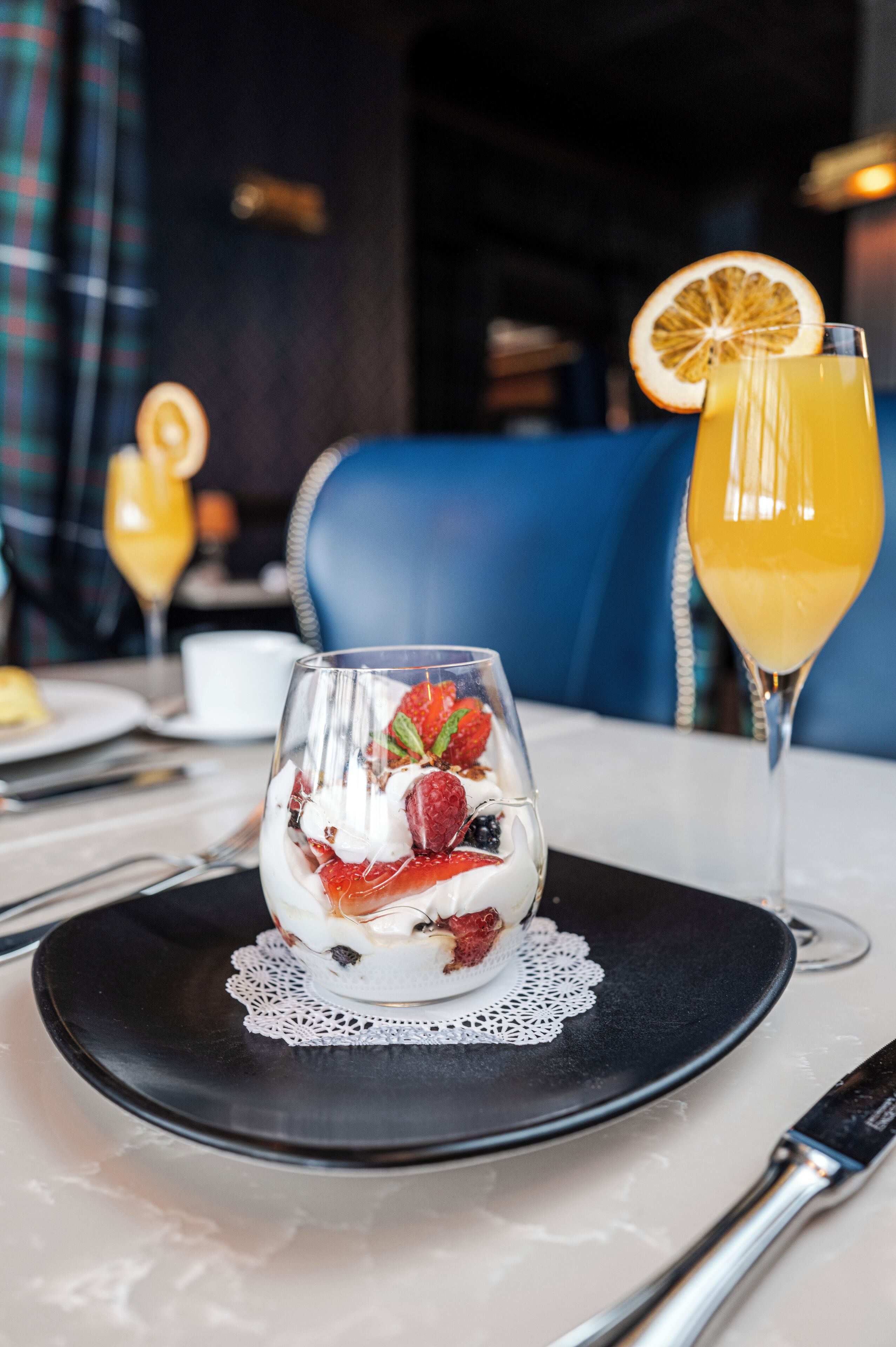 A dessert with strawberries and cream on a black plate, a glass of orange juice, and a blue chair in the background.