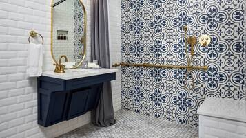 Un baño con pared de ladrillo blanco, lavamanos blanco con detalles dorados, espejo y ducha con pared de azulejos blancos y azules.