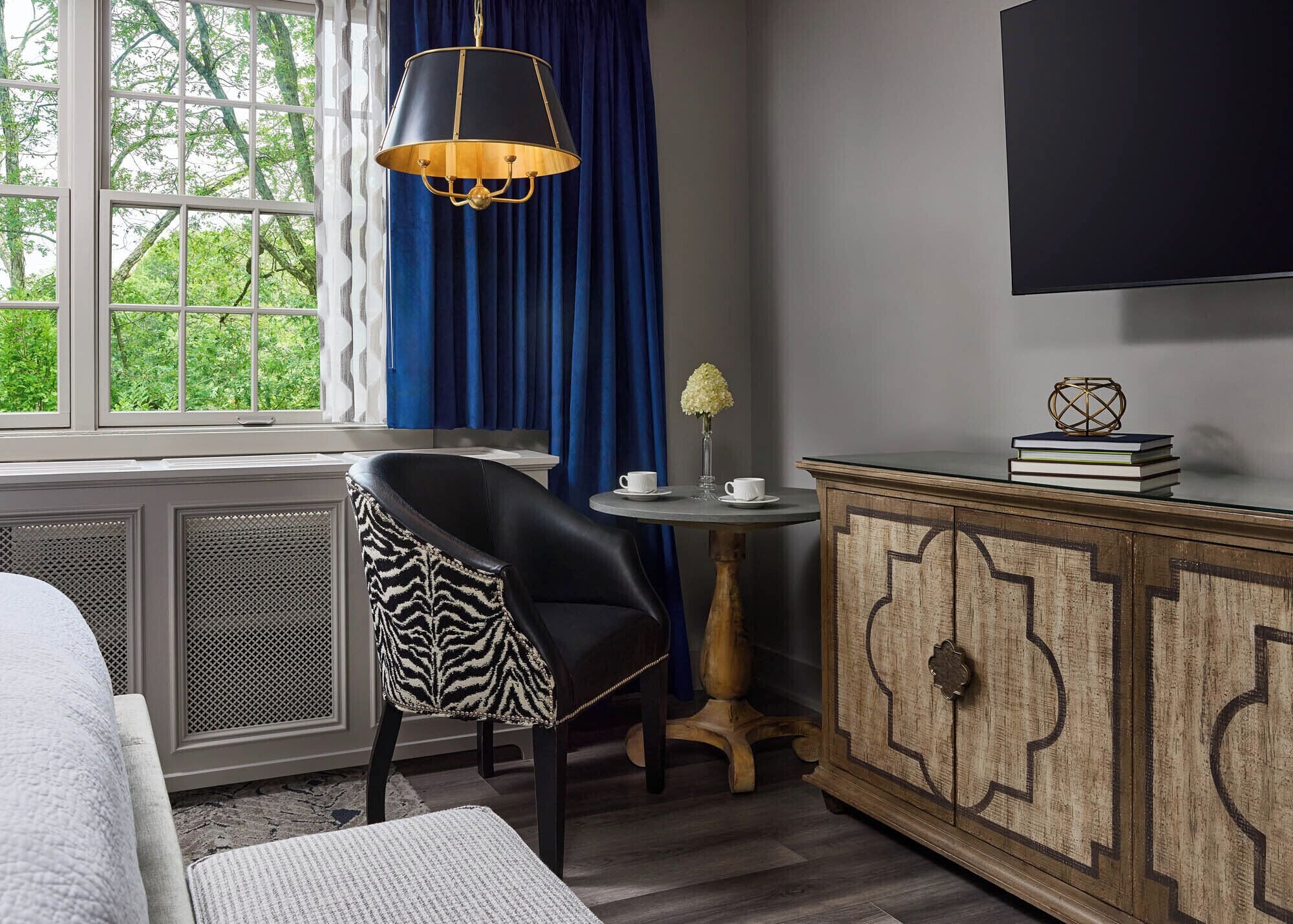 A room with a bed, a zebra-patterned chair, a small round table with a vase, a wooden cabinet, and a hanging lamp.