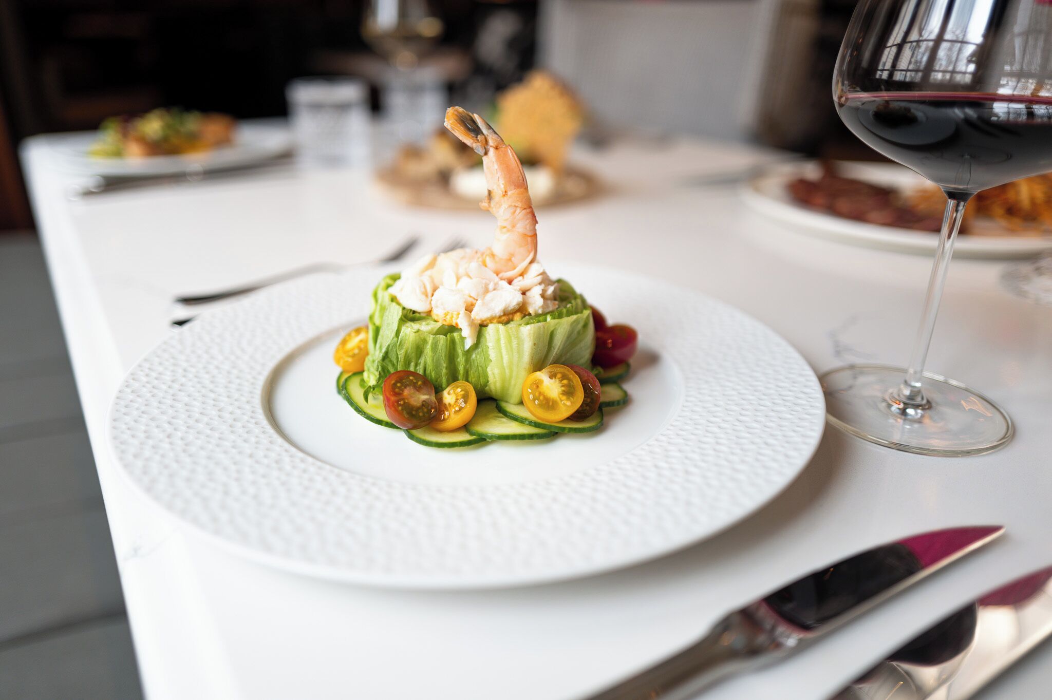 A plate with a shrimp on a bed of lettuce and sliced vegetables, a glass of red wine, and a white tablecloth.