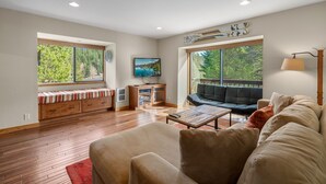 A spacious living room with a sofa, a coffee table, and a TV. There is a window with a view of trees and a balcony with a sofa.