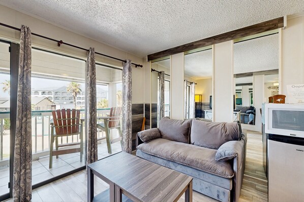 Ein modernes Wohnzimmer mit einer Couch, einem Tisch und einem Fernseher. Es gibt einen Balkon mit einem Sessel und Vorhängen.