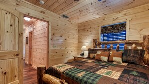 A wooden cabin bedroom with a bed, a nightstand, a lamp, and a window with a view of the forest.