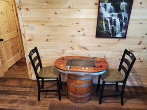 Interior de uma cabana de madeira, com uma mesa e cadeiras rústicas de madeira, um banquinho de metal e um quadro com uma cachoeira na parede.