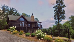 A two-story wooden house with a stone chimney, a black fence, and a gravel driveway.