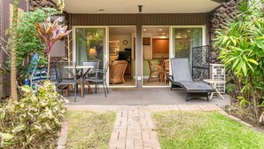A modern building with a covered outdoor area featuring a dining table and chairs, a seating area with a bench, and a small garden with plants. - Hale Hui Kai 102 on Keawakapu Beach (Kihei)