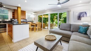 Una cocina moderna con una isla central, un comedor con muebles de madera, un sofá gris con un cojín estampado y un ventilador de techo.