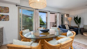 A modern living room with a glass-top dining table surrounded by wicker chairs, a blue sofa, and a large potted plant.