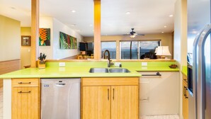 A kitchen with a green countertop, wooden cabinets, a dishwasher, and a view of the ocean through large windows.