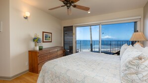 A bedroom with a large bed, a ceiling fan, a dresser, a nightstand, a lamp, and a view of the ocean through a sliding glass door.