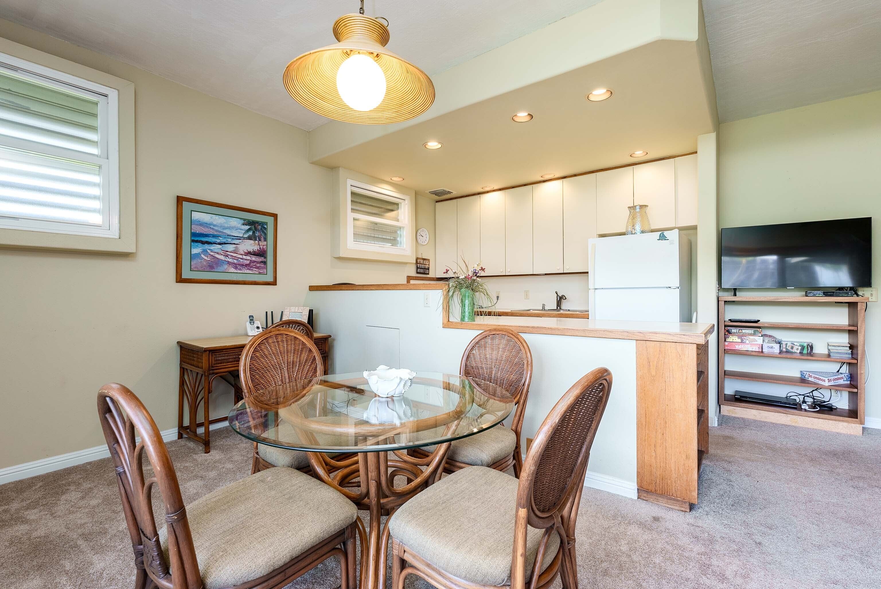 A dining area with a glass table and wicker chairs, a kitchen with white cabinets, a TV on a shelf, and a painting on the wall.