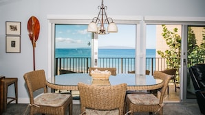 A dining area with wicker furniture, a glass table, and a view of the ocean through a sliding glass door. - Hale Hui Kai 310 Ocean Front Two bedroom / Two bath (Kihei)