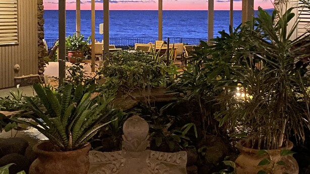 A multi-level indoor space with a view of the ocean at sunset, featuring potted plants and a decorative stone structure.