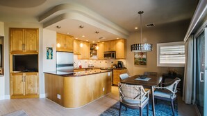A modern kitchen with wooden cabinets, a central island, and a dining area with wicker chairs. - Hale Hui Kai 308 OCEAN VIEW (Kihei)