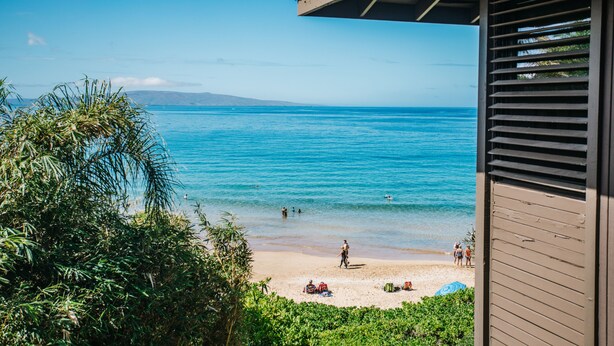 A beach view with clear blue water, a sandy shore, and a building with a sloped roof. - Hale Hui Kai 308 OCEAN VIEW (Kihei)