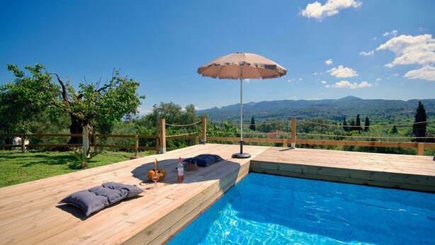 Un terrazzo in legno con piscina, ombrellone e vista sulle montagne.