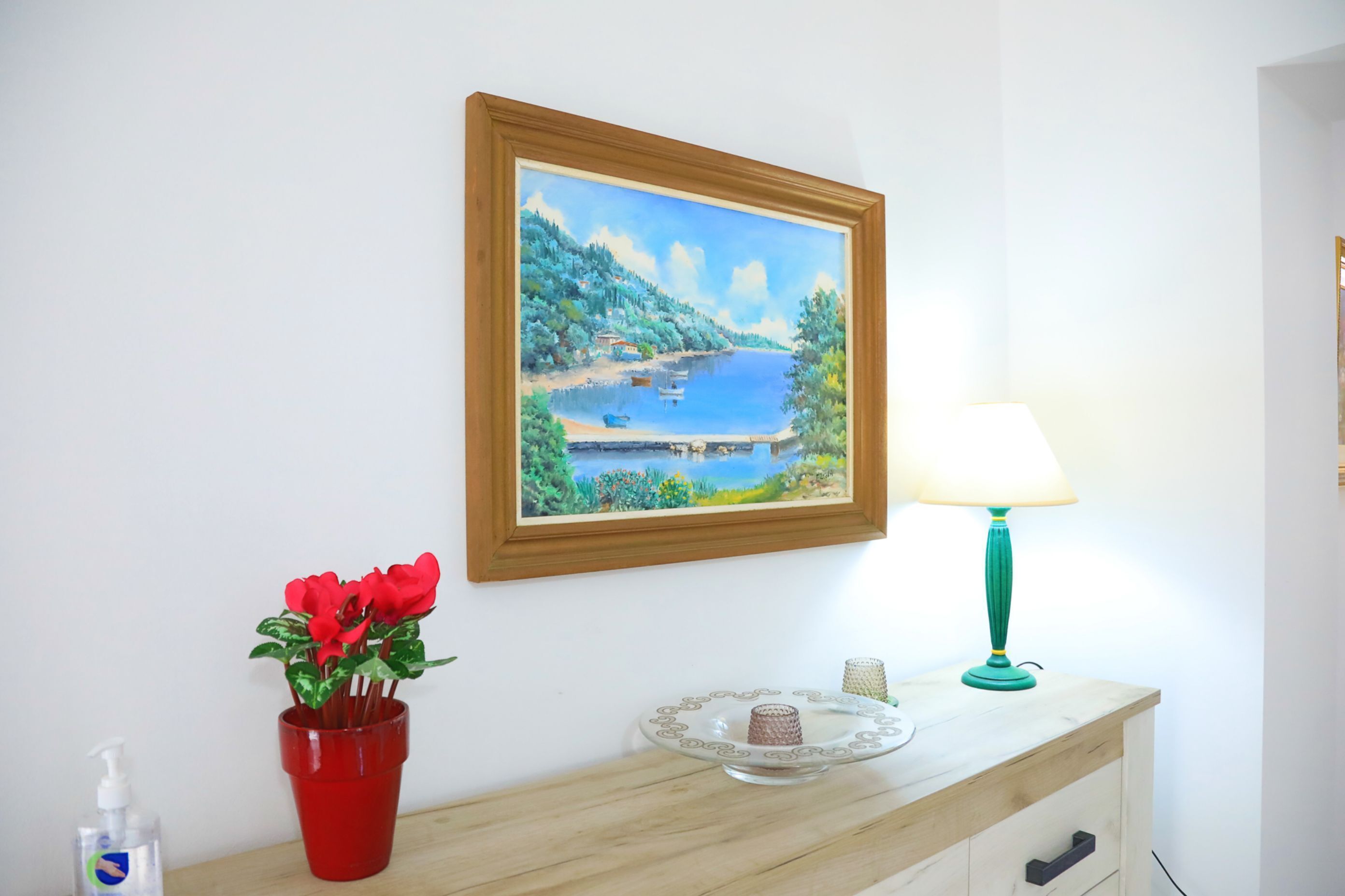 Ein Zimmer mit einem Holzkoffer, einem gerahmten Gemälde mit Seeblick, einer roten Blumenvase, einer blaugrünen Lampe und einem kleinen Tisch mit dekorativer Platte und Kerze.