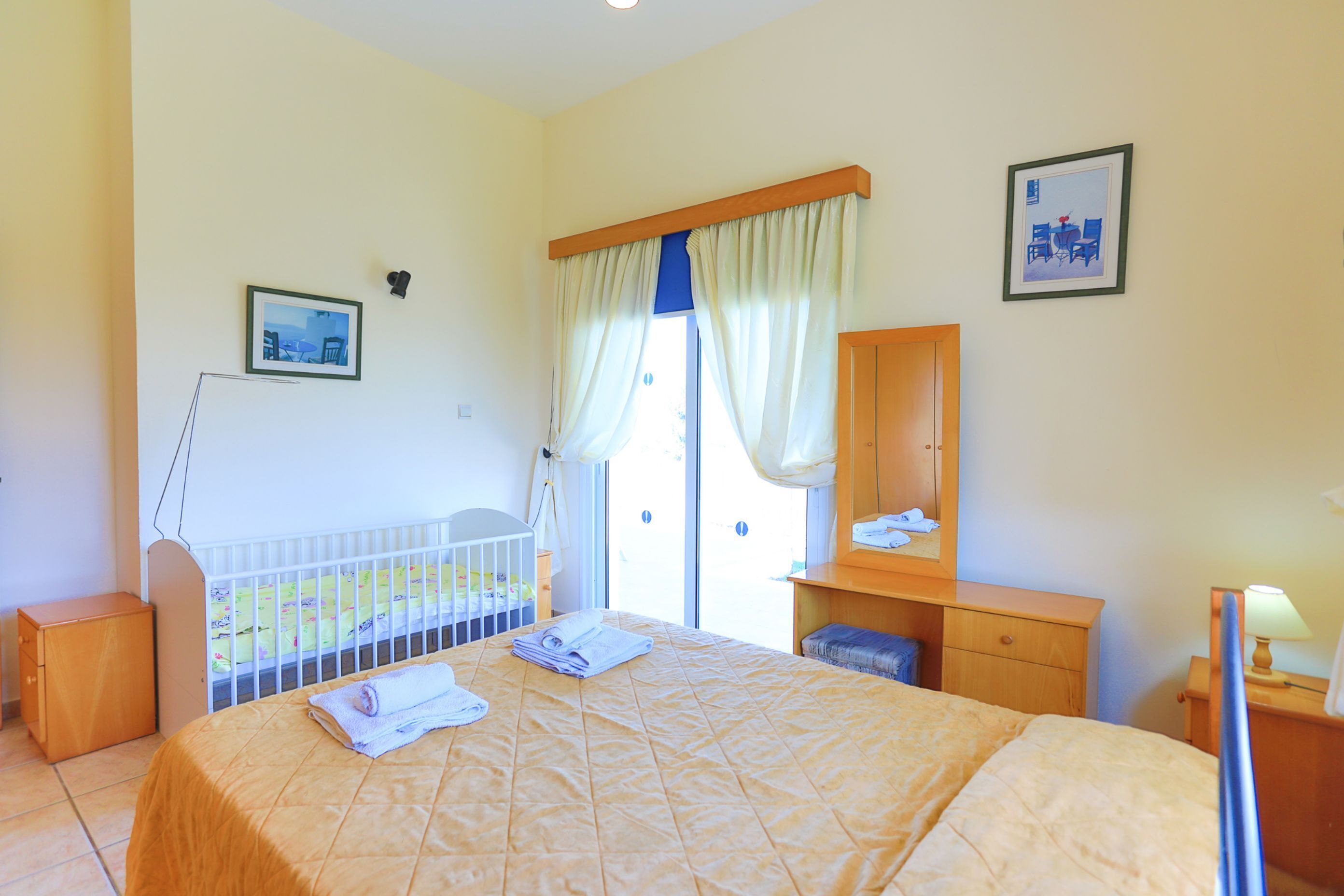 A hotel room with a bed, a desk, a chair, a small table, a crib, and a window with curtains.