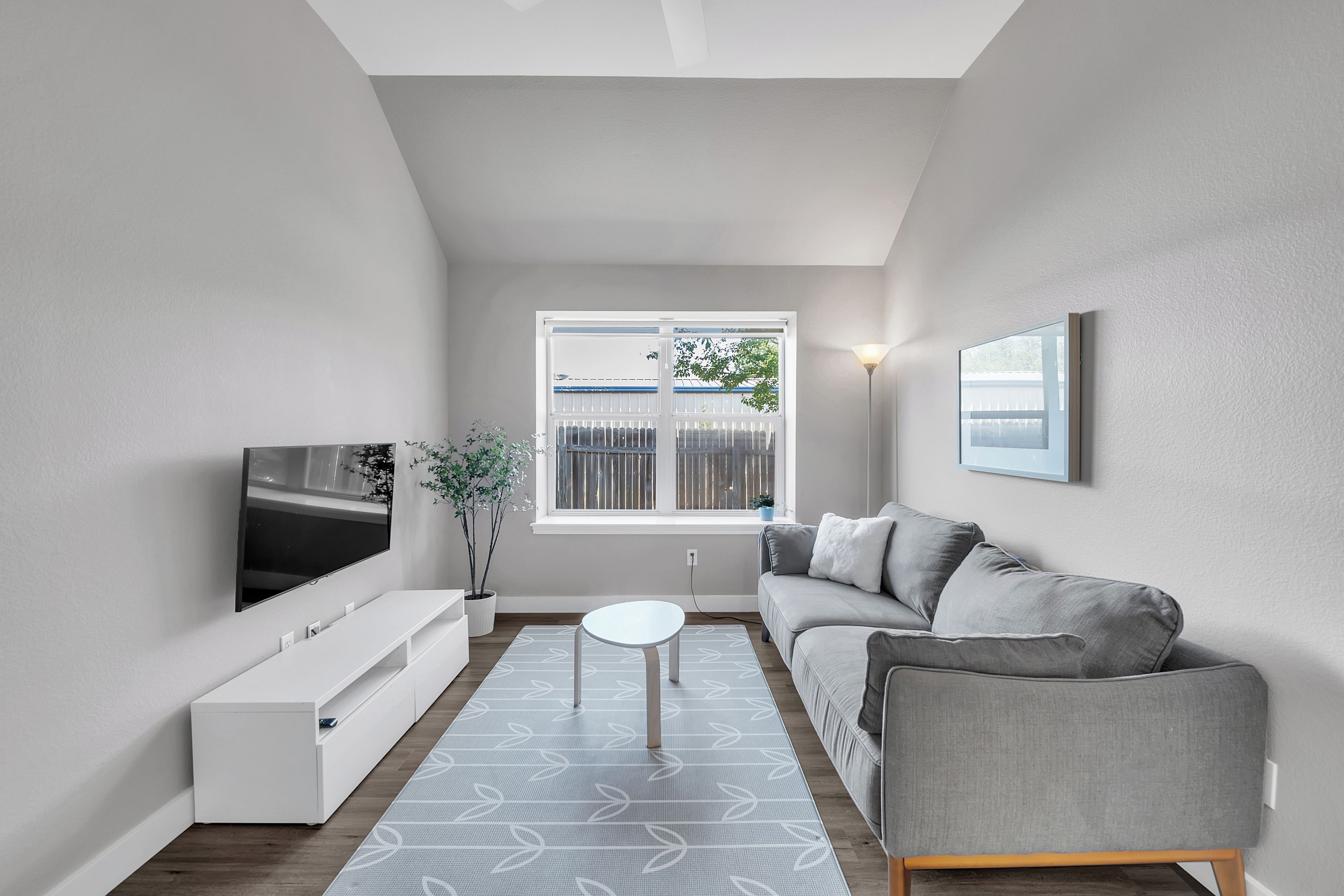 A modern living room with a grey sofa, a flat-screen TV mounted on the wall, a white coffee table, and a window with a view of the outside.
