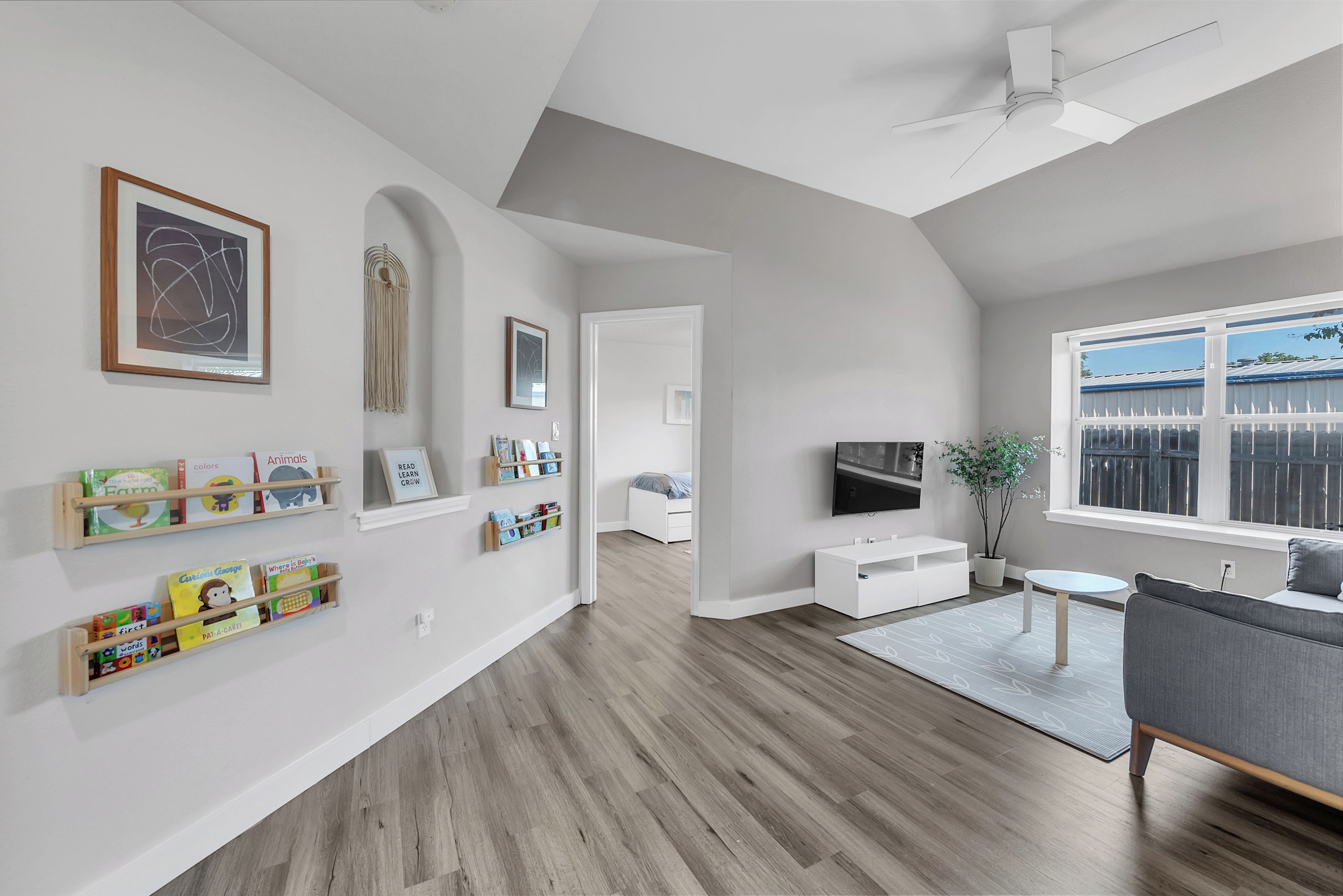A modern living room with a wooden floor, a grey sofa, a TV, and a bookshelf with children's books.