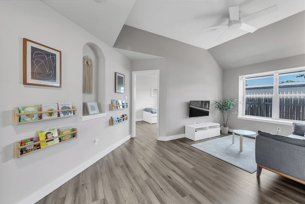 A modern living room with a wooden floor, a grey sofa, a TV, and a bookshelf with children's books.
