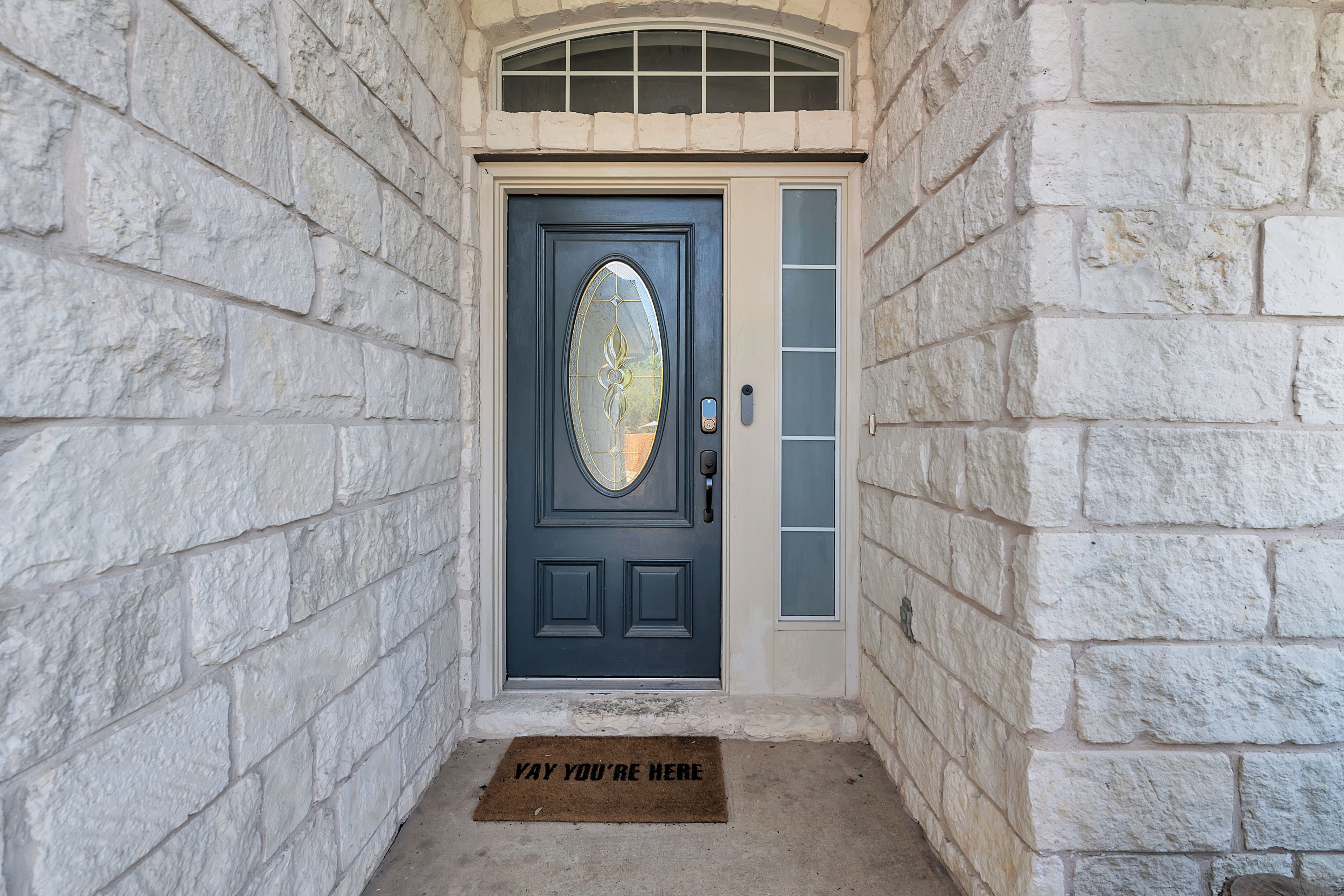 A stone-walled entrance with a dark door featuring a glass panel and a doormat with text.